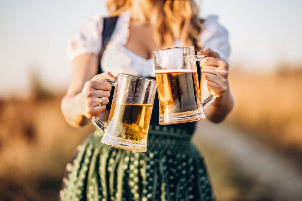 Close-up photo of blonde in dirndl, traditional festival dress, holding two mugs of beer in her hands outdoors. Oktoberfest, St. Patrick’s day, international beer day concept.