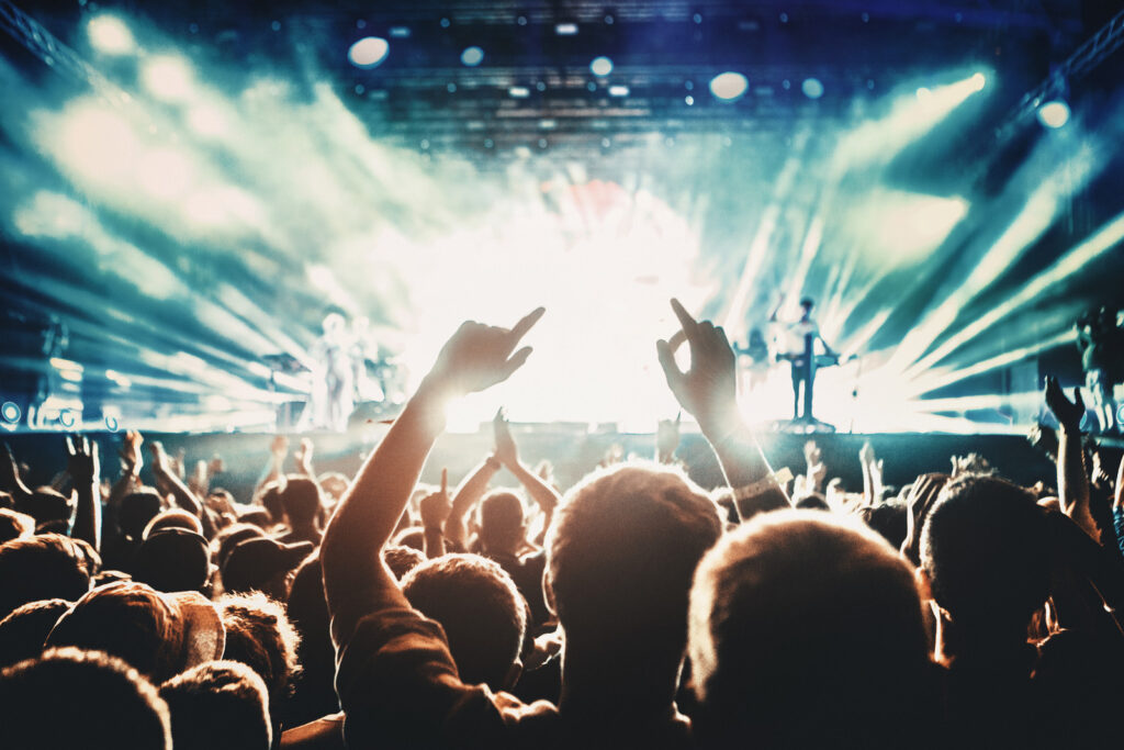 Rear view of people cheering at concert during music festival.Large group of unrecognizable adults with their arms raised.Some of them holding beer cans,some are taping the show with phones.There's a stage and a band performing in background,out of focus.Stage is toned cyan blue.