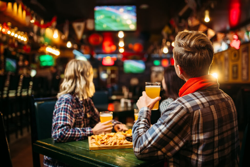 Football fans watching match and drinks beer at the table in sports bar. Tv broadcasting, young friends leisures in pub